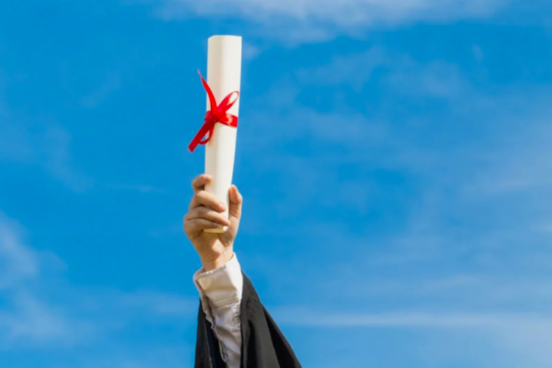 Mavi gökyüzüne doğru uzatılmış, kırmızı kurdeleyle bağlanmış bir diploma rulosu, mezuniyet ve akademik başarıyı simgeleyen görsel.