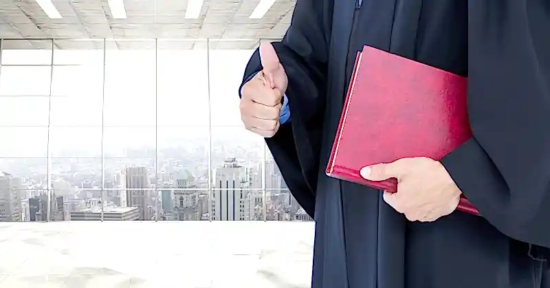 Akademik cübbe giymiş kişi kitap tutuyor ve başparmağını kaldırıyor – akademik kadro ilanlarına itiraz ve dava süreçleri hakkında görsel anlatım.