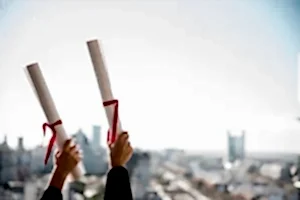 Şehir manzarası ve açık gökyüzü önünde, başarıyla tamamlanan hukuki eğitimi simgeleyen iki kırmızı kurdeleli diploma rulosunu havaya kaldıran eller.