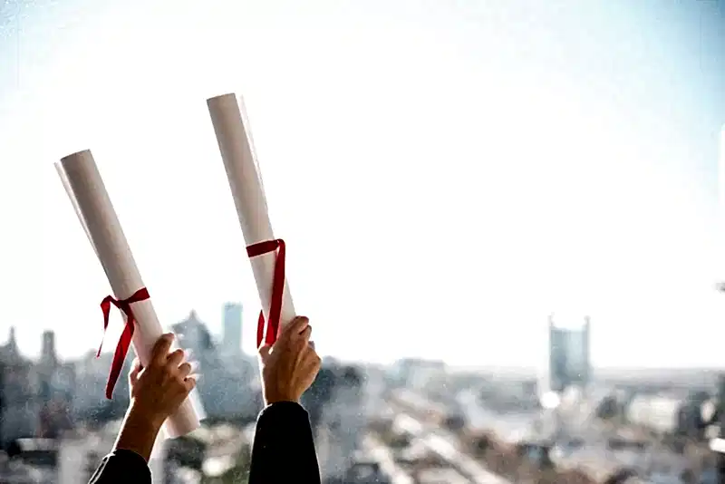 Şehir manzarası ve açık gökyüzü önünde, başarıyla tamamlanan hukuki eğitimi simgeleyen iki kırmızı kurdeleli diploma rulosunu havaya kaldıran eller.
