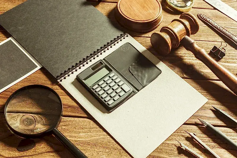 Ahşap bir masa üzerinde, hukuki muhasebeyi ve finansal davaları temsil eden bir tokmak, hesap makinesi, büyüteç ve not defteri.