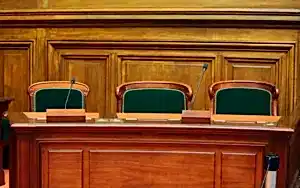 Boş bir mahkeme salonunda, hakimin veya jürinin oturduğu ahşap kürsü ve yeşil sandalyelerle, hukuki duruşmaları temsil eden görsel.