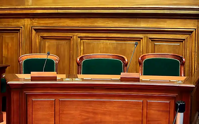 Boş bir mahkeme salonunda, hakimin veya jürinin oturduğu ahşap kürsü ve yeşil sandalyelerle, hukuki duruşmaları temsil eden görsel.