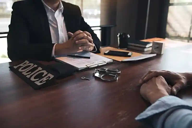 Bir polis memurunun bir şüpheliyi sorguladığı veya ifadesini aldığı anı gösteren, masada "POLICE" yazılı kol bandı ve kelepçeler bulunan görsel.