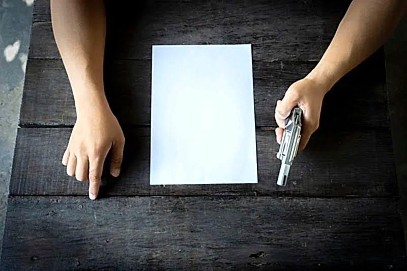 Karanlık ahşap bir masa üzerinde, boş bir kağıda doğru işaret eden bir el ve yanında duran bir tabancayla ceza hukuku davalarındaki ciddi durumu temsil eden görsel.